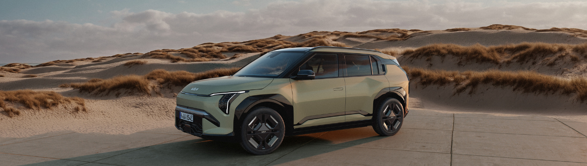 Kia EV3 | Das Bild zeigt einen hellblauen SUV auf einem Parkplatz vor Sanddünen.