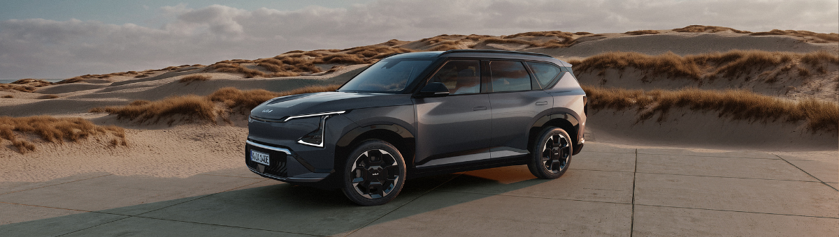 Kia EV5 | Das Bild zeigt einen hellblauen SUV auf einem Parkplatz vor Sanddünen.